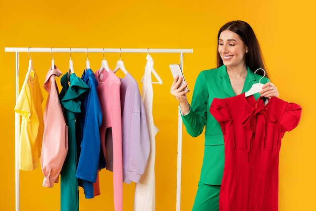 Senhora sorridente feliz usando smartphone e segurando vestido de mulher pedindo roupas on-line no fundo amarelo do aplicativo móvel