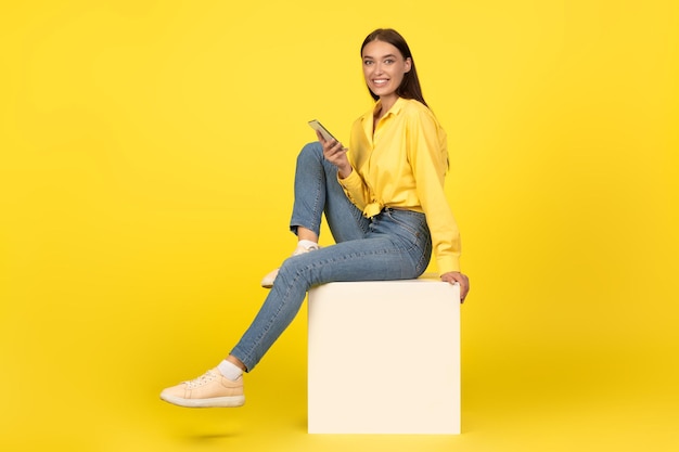 Senhora usando o celular sorrindo para a câmera sentada sobre fundo amarelo