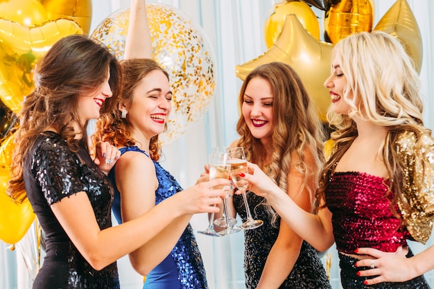 Senhoras em vestidos de lantejoulas. Garotas felizes tilintando de taças com champanhe, rindo, se divertindo.