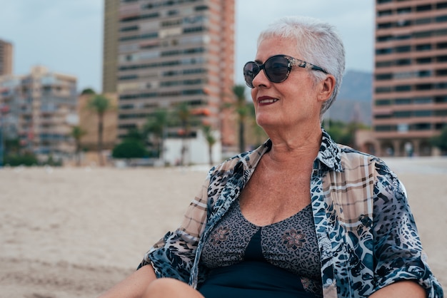 Senior anciana en la playa con gafas de sol cabello gris sentado en la playa