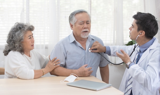 Senior anciano asiático anciano con máscara en acostado en el sofá mientras joven médico caucásico sentarse en la rodilla comprobar su latido