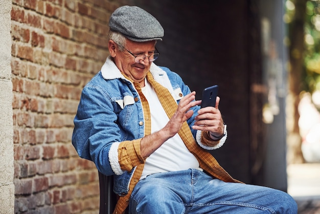 Sênior elegante em roupas da moda e de óculos se senta na cadeira e usa o telefone.