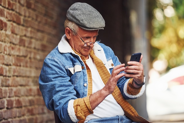 Sênior elegante em roupas da moda e de óculos se senta na cadeira e usa o telefone.