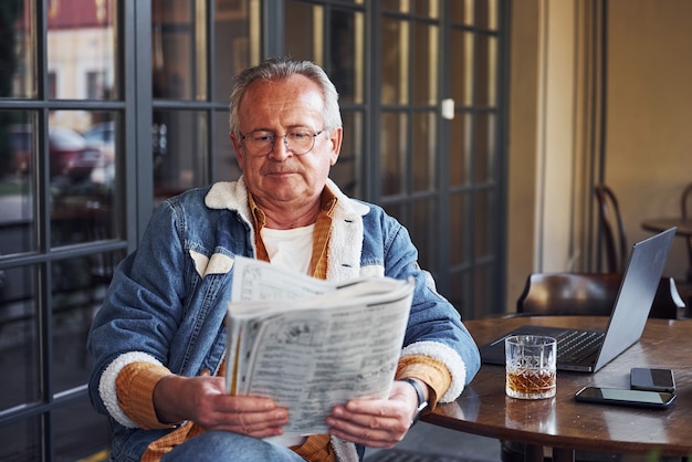 Sênior elegante em roupas da moda e de óculos se senta no café e lê o jornal.