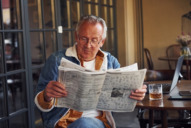 Foto sênior elegante em roupas da moda e de óculos se senta no café e lê o jornal.