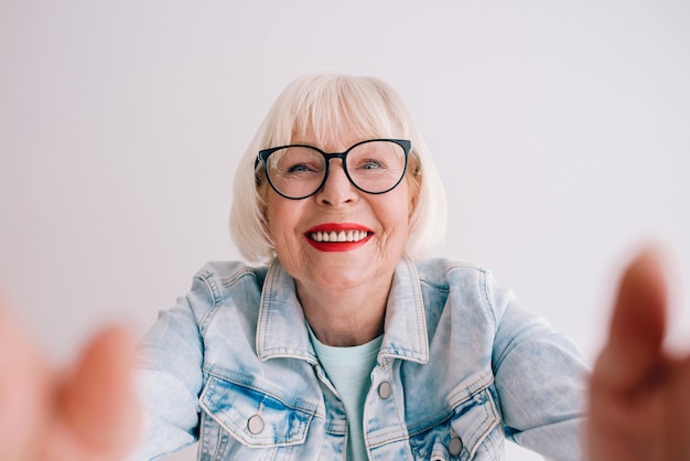 Senior elegante mujer con cabello gris y gafas y chaqueta de jeans abrazándote o haciendo selfie