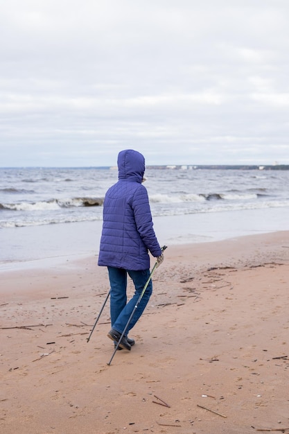 Senior Frau im Ruhestand Wandern am Meer auf dem Sand Aktive Fitness und gesunde Lebensweise Weibliches Training am Strand Nordic Walking mit Stöcken Herbstsaison am Meer Sportliches Paar