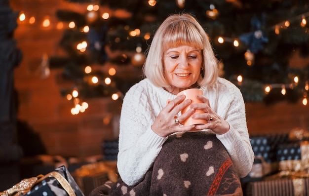 Senior fröhliche Frau sitzt mit einer Tasse Getränk im Zimmer mit Weihnachtsgeschenkboxen.