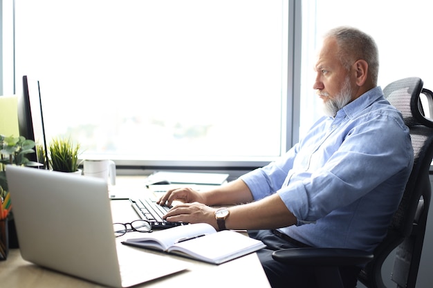 Senior Geschäftsmann mit einem stylischen kurzen Bart, der an seinem Schreibtisch an einem Laptop arbeitet.