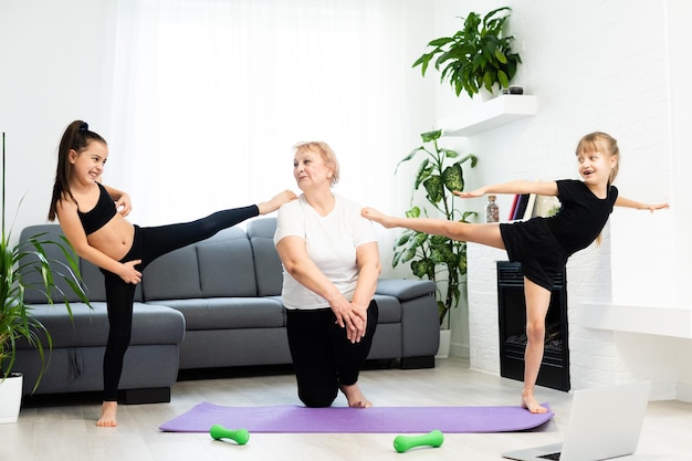 senior großmutter training yoga mit enkelinnen im vorschulalter. Sie sitzen auf einer Matte in einem gemütlichen Zuhause. Sport, Elternschaft und People-Konzept.