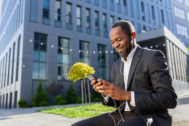 Senior hombre afroamericano sonriente hombre de negocios en traje sentado en un banco fuera del centro de oficinas