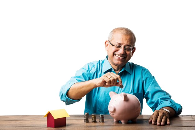 Senior hombre asiático indio y concepto de ahorro - con hucha, billetes, modelo de casa 3d, ventilador de dinero, pila de monedas. sentado a la mesa