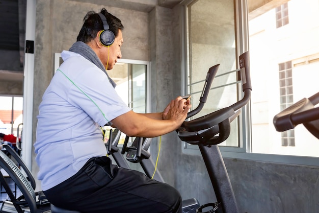 Senior hombre asiático en ropa deportiva escuchar música y entrenamiento ciclismo cardio en el gimnasio.