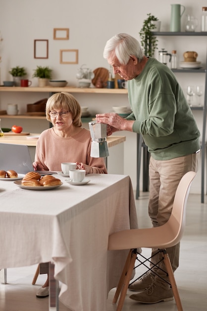 Senior hombre cuidando a su esposa vertiendo té caliente durante el desayuno mientras ella está sentada a la mesa con un portátil
