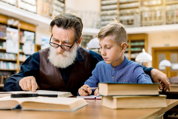 Senior hombre guapo, leyendo un libro en voz alta a su nieto o estudiante, que lo escucha con atención y toma notas. Abuelo y nieto en biblioteca