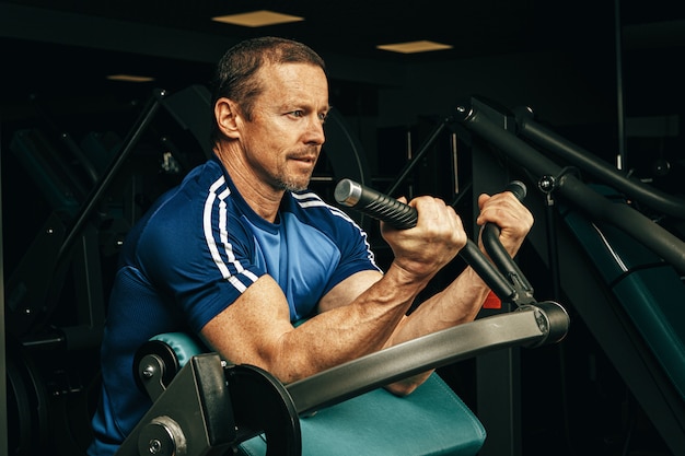 Senior hombre haciendo ejercicios para brazos en una máquina de entrenamiento en gimnasio