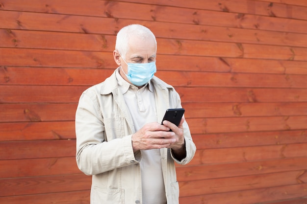 Senior hombre con mascarilla médica usando el teléfono para buscar noticias