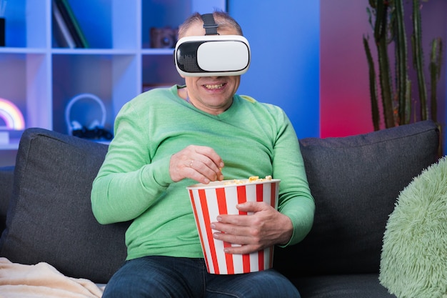 Foto senior hombre sentado en el sofá y sosteniendo palomitas de maíz, con gafas de realidad virtual y levanta su mano para seleccionar con pantalla vr.