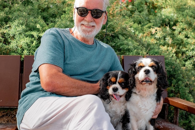 Senior hombre sonriente con gafas de sol sentado en un banco con sus dos perros cavalier king charles spaniel