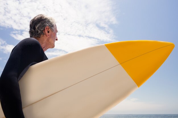 Senior hombre en traje de neopreno con una tabla de surf y mirando a una distancia