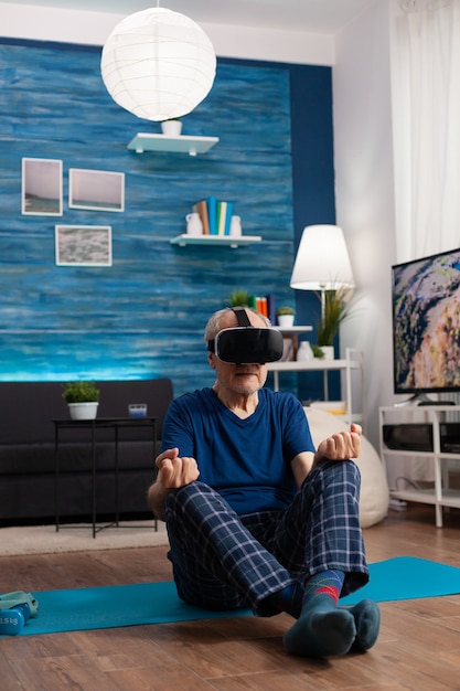 Foto senior hombre vestido con casco de realidad virtual meditando mientras está sentado en posición de loto en la estera de yoga en la sala de estar durante pilates