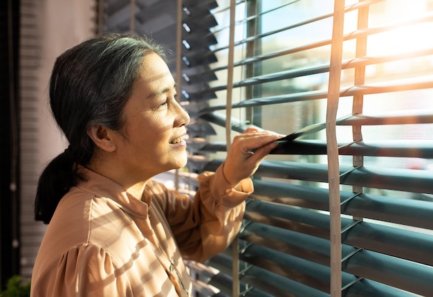 Senior Ältere Frau schaut abends auf das Fenster Großmutter wartet geduldig auf die nach Hause kommende Person mit trauriger, einsamer Depression Asiatische 70er-Jahre-Frau wartet mit ernster Sorge und fühlt sich kopiert