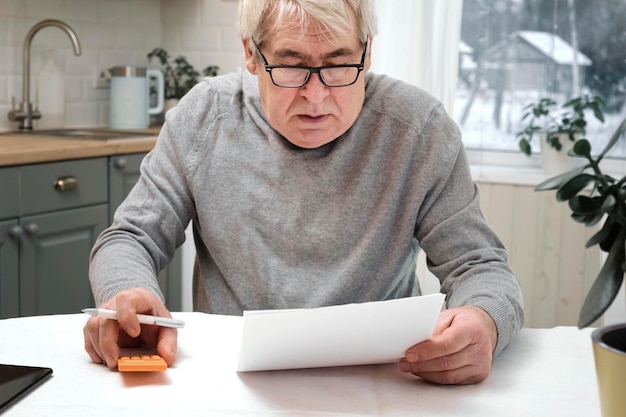 Senior Mann, der die Berechnung der persönlichen Finanzen zu Hause überprüft und sich durch Geldprobleme gestresst fühlt. Besorgte ältere Männer erhalten eine offizielle Bankbenachrichtigung über unbezahlte Schulden oder Steuern und fühlen sich besorgt