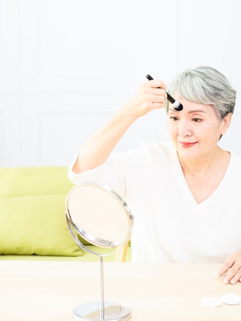 Senior mujer asiática aplicando base en la mejilla con un pincel de maquillaje mientras está sentada sola frente a un espejo.