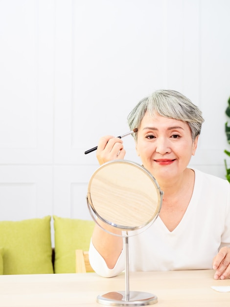 Senior mujer asiática aplicando base en la mejilla con un pincel de maquillaje mientras está sentada sola frente a un espejo.