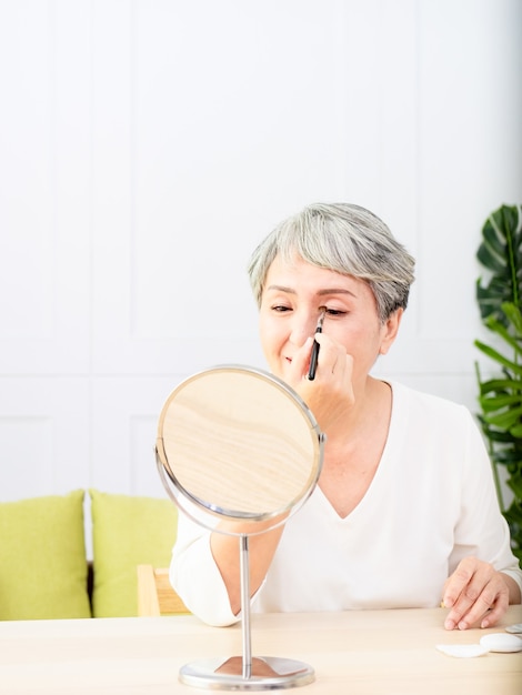 Senior mujer asiática aplicando base en la mejilla con un pincel de maquillaje mientras está sentada sola frente a un espejo.