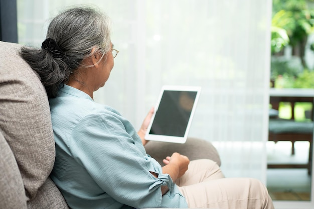 Senior mujer asiática con gafas y relajarse en casa en un sofá y usar una tableta para leer noticias y libros electrónicos. Concepto de tecnología para personas mayores.