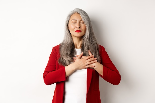 Senior mujer asiática con labios rojos y chaqueta, ojos cerrados y tomados de la mano en el corazón agradecido, sintiéndose nostálgico, soñando con algo, de pie sobre fondo blanco.