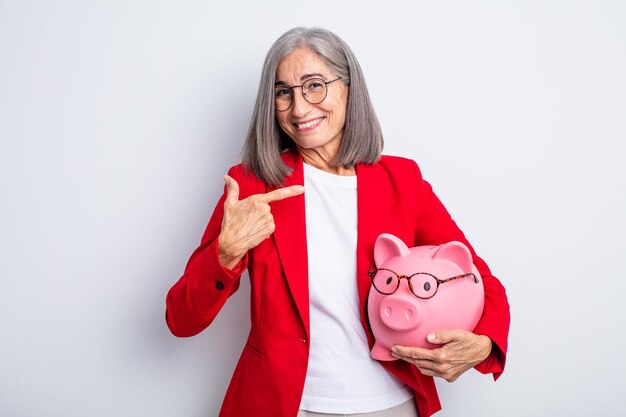 Senior mujer bonita sonriendo alegremente sintiéndose feliz y apuntando al concepto de alcancía lateral