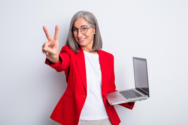 Senior mujer bonita sonriendo y luciendo feliz gesticulando victoria o concepto de negocio y computadora portátil de paz