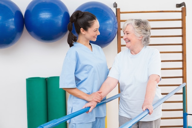 Senior mujer caminando con barras paralelas con terapeuta