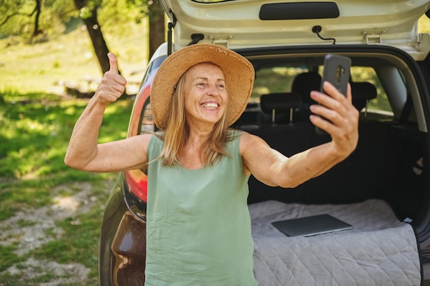 Senior mujer feliz activa hace selfie con teléfono en el maletero del coche o arranque en camper soleado parque de verano