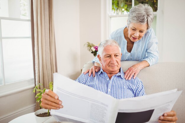 Senior mujer hablando con senior hombre leyendo el periódico en la sala de estar