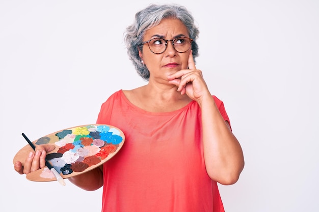 Senior mujer hispana de cabello gris sosteniendo pincel y paleta cara seria pensando en la pregunta con la mano en la barbilla pensativa sobre una idea confusa