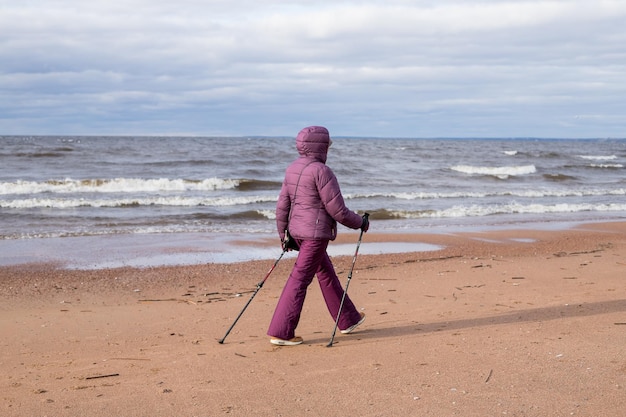 Senior mujer jubilada haciendo senderismo en la playa en la arena Fitness activo y estilo de vida saludable Entrenamiento femenino en la playa Marcha nórdica con bastones Temporada de otoño en la playa Pareja deportiva