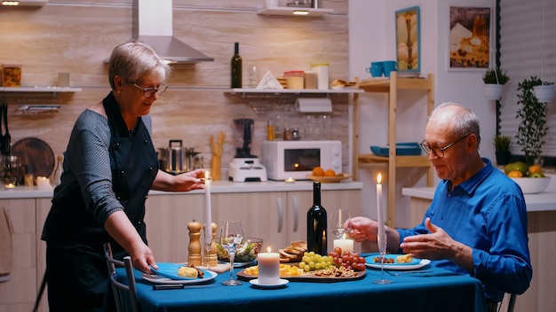 Senior mujer madura jubilada que sirve la cena en el comedor. Pareja de ancianos hablando, sentados a la mesa en la cocina, disfrutando de la comida, celebrando su aniversario con comida sana.