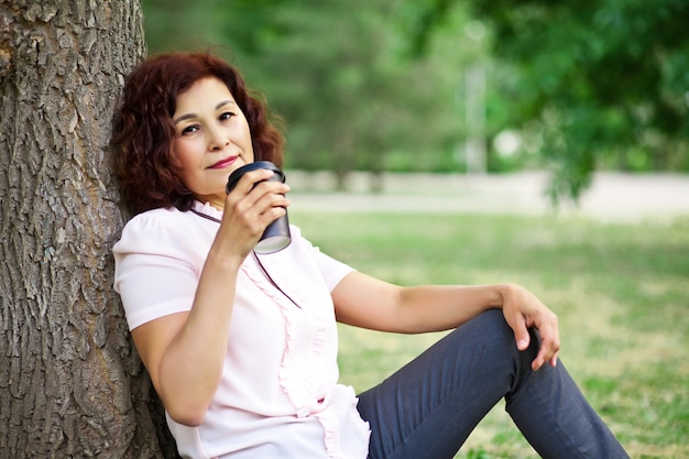 Senior mujer de mediana edad sentada en un parque al aire libre y bebiendo café de un vaso de papel