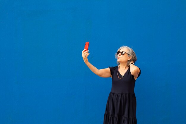 Senior mujer de pelo gris tomando un selfie con un smartphone rojo contra una pared azul