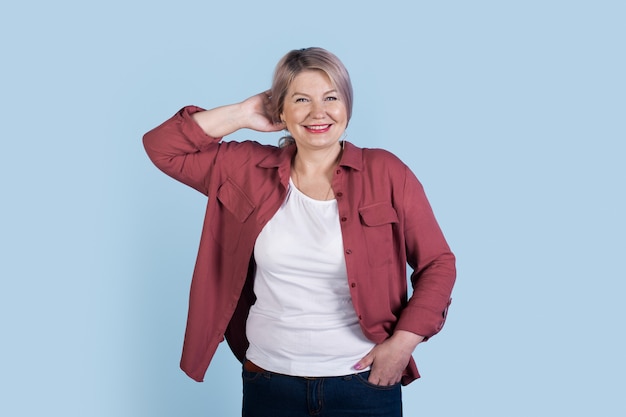 Senior mujer rubia está sonriendo a la cámara con una camisa y posando en una pared azul del estudio