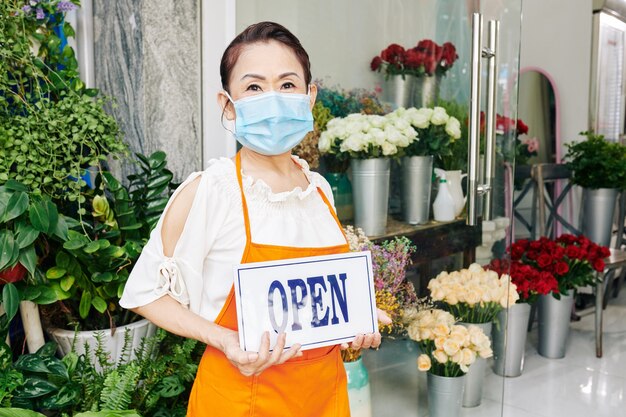 Senior mujer vietnamita en máscara médica mostrando signo abierto cuando está de pie en la florería