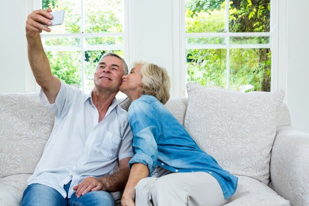 Sênior mulher beijando enquanto homem tomando selfie na sala de estar