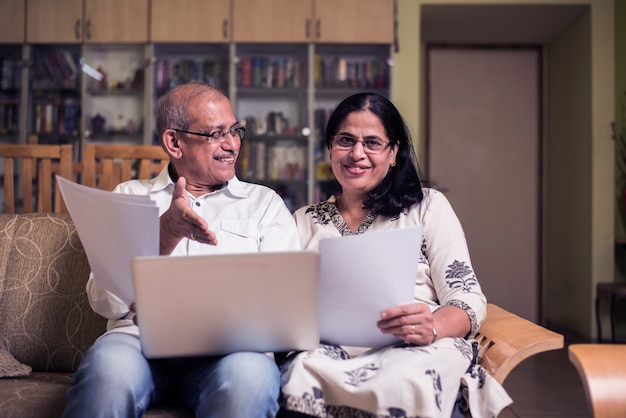 Senior pareja asiática india contabilidad, haciendo finanzas para el hogar y verificando facturas con computadora portátil, calculadora y dinero también con hucha mientras está sentado en el sofá, sofá o mesa en casa