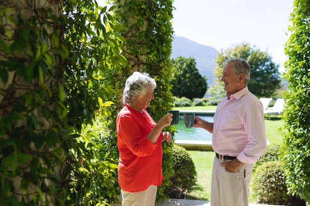 Senior pareja caucásica sonriendo y sosteniendo tazas en un jardín soleado. concepto de retiro, jubilación y estilo de vida senior feliz.