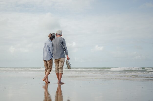 Senior pareja cogidos de la mano y caminando por la playa, la otra mano de los dos estaba sosteniendo un par de zapatos. Plan de seguro de vida en el concepto de jubilación.