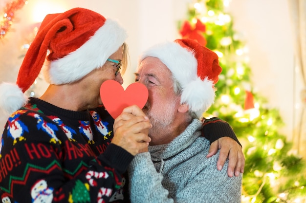 Senior pareja de jubilados mirándose a los ojos sosteniendo un corazón rojo de papel con gorro de Papá Noel