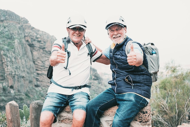 Senior sonriente pareja de hombres excursionistas con cascos y mochilas descansando en la cima de la montaña